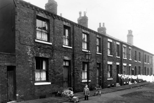 A row of back-to-back terraced houses with a yard on the left built to house the shared outside toilets in February 1959. A child stands in the street while toy prams and two tricycles lay diacarded in the road. Clothes hang out on lines stretched across the street.