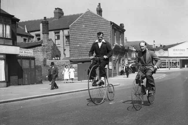 Two men riding a bone-shaker bicycle and a penny farthing on Vicarage lane, Blackpool, with the Oxford Garage on Waterloo Road in the background. The photograph was taken in the 1950s when the bicycles were already quite old
