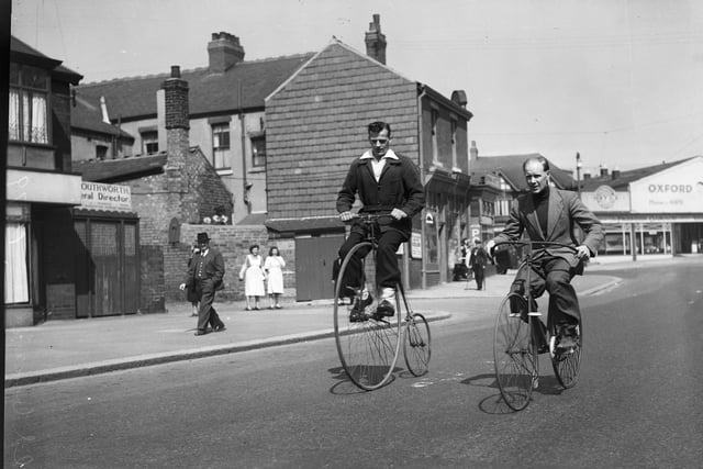 Two men riding a bone-shaker bicycle and a penny farthing on Vicarage lane, Blackpool, with the Oxford Garage on Waterloo Road in the background, 1950s