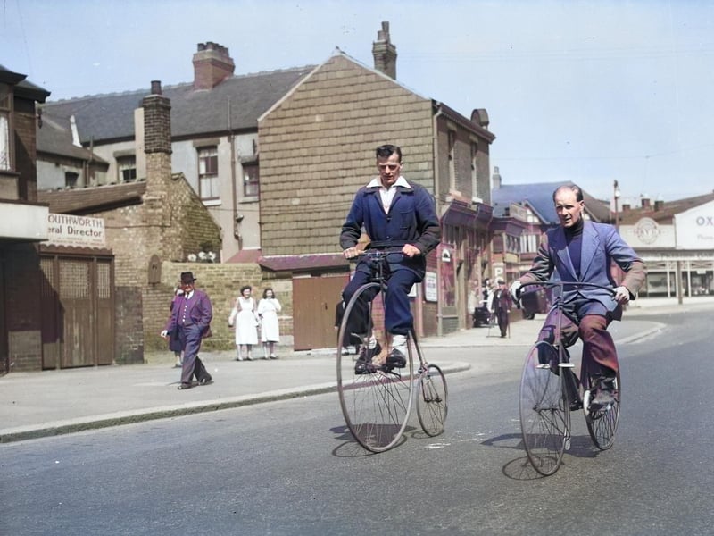 Two men riding a bone-shaker bicycle and a penny farthing on Vicarage lane, Blackpool, with the Oxford Garage on Waterloo Road in the background. Their clothing is striking in colour. Probably the 1950s