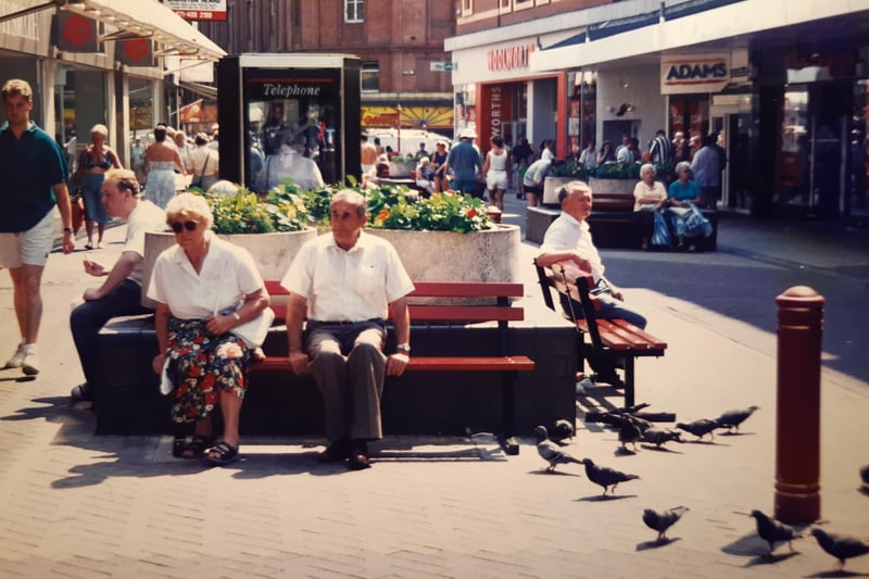 A telephone box, summer bedding plants and red street furniture for this 1995 scene. Adams children's wear, Miss Selfridge, Woolworths and WH Smith can be seen